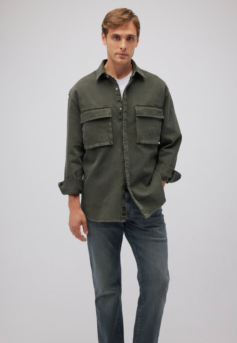 Dunkelgrüne Denim-Hemd mit zwei Fronttaschen, Knopfleiste und hochgerollten Ärmeln, kombiniert mit blauen Jeans. Schlichter Hintergrund.