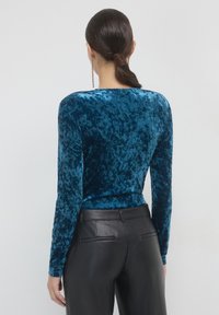 Top a maniche lunghe in velluto teal con una texture liscia, dotato di scollatura a girocollo e una silhouette aderente, abbinato a pantaloni in pelle nera.