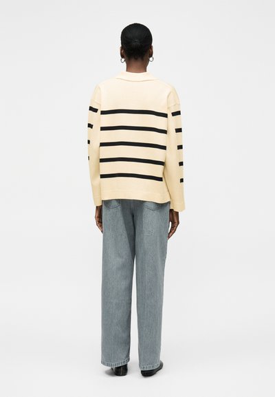 Άτομο με σκούρο δέρμα και μαλλιά σε κότσο, φοράει κρεμ πουλόβερ με μαύρες οριζόντιες ρίγες και άνετο ανοιχτό μπλε τζιν, στέκεται γυρισμένο με την πλάτη.