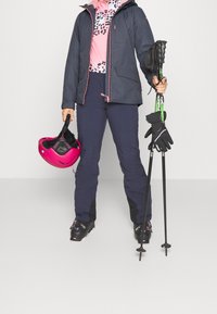 Strój narciarski z granatową kurtką, różową bluzą z leopardzim nadrukiem, granatowymi spodniami, czarnymi rękawicami, różowym kaskiem i kijami narciarskimi.