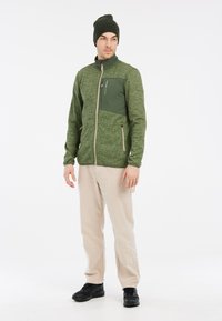 Groene fleecejack met een gestructureerd oppervlak, voorrits en twee ritszakken. Gepaard met beige broeken en zwarte schoenen, afgemaakt met een groene muts.