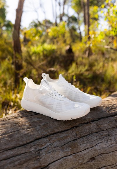 Chaussures de sport blanches avec un dessus en maille respirante, design texturé, fermeture à lacets et semelle renforcée, posées sur un tronc en bois.