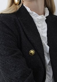 Czarna blazer z fakturą, z złotym guzikiem, nałożona na białą bluzkę z falbankami. Skupienie na detalach tkaniny i wzorze guzika.