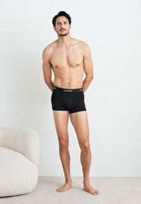 Zwarte katoenen boxershorts met een logo-elastische tailleband, model staat blootsvoets, kort haar, effen achtergrond.