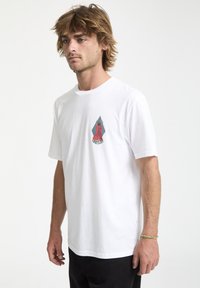 Hvid kortærmet t-shirt med et farverigt geometrisk grafik på venstre bryst. Glat bomuldsstruktur, afslappet pasform. Sorte bukser på.