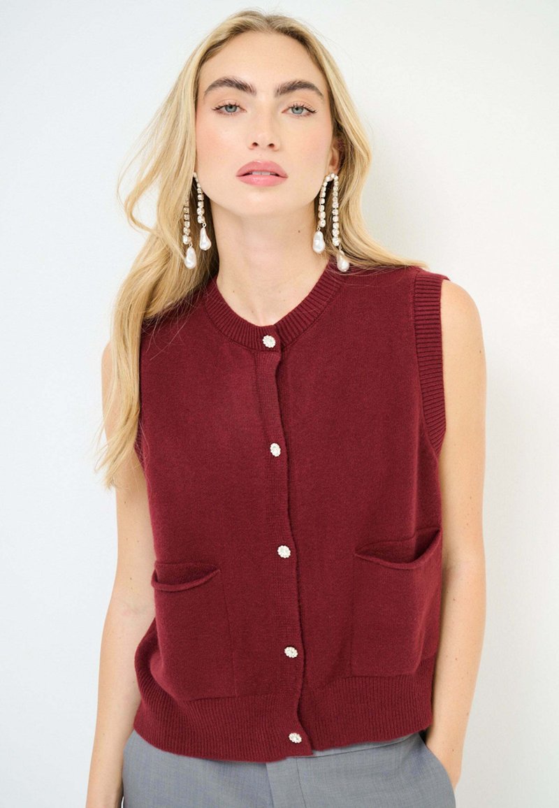 Cardigan senza maniche bordeaux con scollo rotondo, due tasche frontali e dettagli in bottoni bianchi, realizzato in morbido tessuto a maglia.