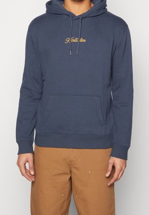 Mężczyzna w granatowej bluzie z kapturem z żółtym logo „Hollister” i jasnobrązowych spodniach, stojący na jednolitym tle.
