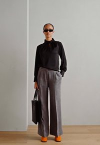 Schwarze Bluse mit Schlüsselaugen-Detail, graue weit geschnittene Hose, orangefarbene Absatzschuhe und eine schwarze Tote-Tasche; glatte Texturen und klare Linien.