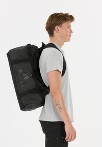 Musta reppu, jossa on säädettävät hihnat, valmistettu vedenkestävästä materiaalista. Repussa on logo ja vahvistetut saumat, ja se on suunniteltu kestäväksi käytössä.