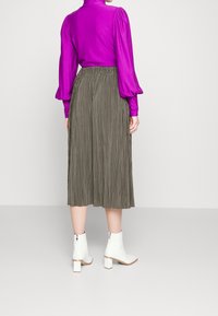 Kvinna klädd i en ljuslila långärmad blus, olivgrön plisserad midikjol och vita ankelstövlar som står mot en neutral bakgrund.