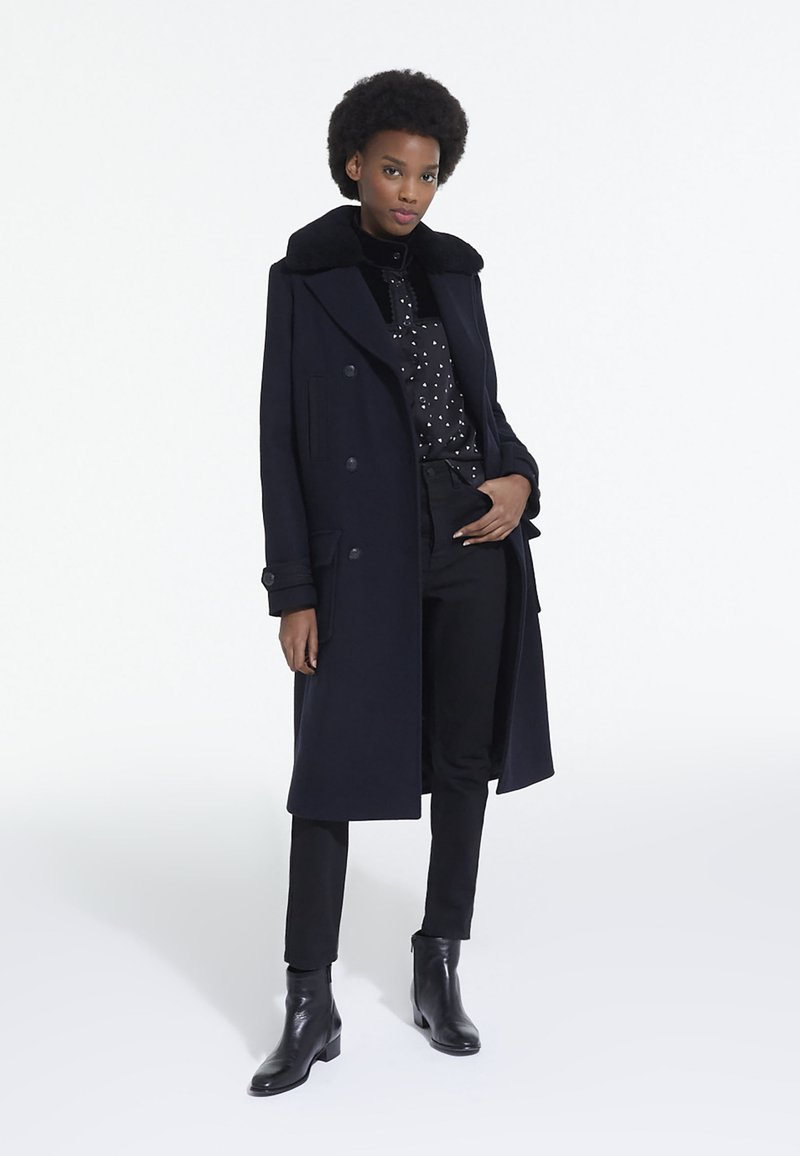 Manteau long bleu marine avec un col doux noir, design croisé, associé à un haut à pois, un pantalon noir et des bottines. Texture lisse.