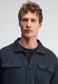 Dunkelgrau wollene Hemdjacke mit Kragen, Vordertaschen und schwarzen Knöpfen. Weiche Textur mit einem leicht strukturierten Schnitt.