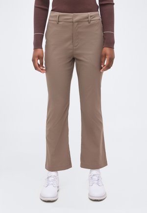 Osoba oblečená v rovnom taupovom nohaviciach, hnedom tričku s dlhým rukávom a bielych teniskách, stojaca pred jednofarebným svetlým pozadím.