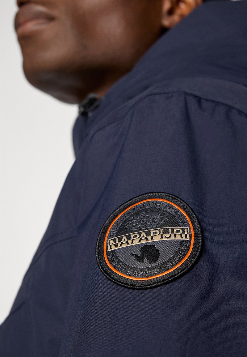 Veste bleu marine à la surface texturée, avec un logo brodé rond noir et orange sur l'épaule gauche.