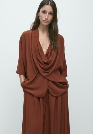 Kobieta o długich brązowych włosach nosi luźną, przewieszoną rdzawo-brązową bluzkę z głębokim dekoltem oraz dopasowane szerokie spodnie, ręce w kieszeniach, neutralne tło.