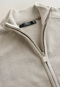 Beige zip-up tröja med ribbat krage och strukturerad stickning. Har en slät dragkedja och en märkesetikett inuti. Mjuk material.