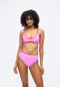 Göndör hajú nő fekete, téglalap alakú napszemüveget visel, rózsaszín bikinit, amelynek piros szegélye van, és egy világos, egyszínű háttér előtt áll.