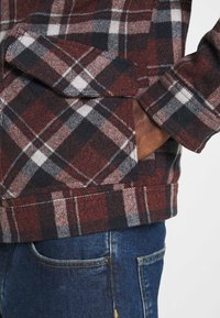 Camicia di flanella a quadri rossi e neri con finitura testurizzata, caratterizzata da una tasca laterale e un polsino orlato, abbinata a jeans di denim blu.