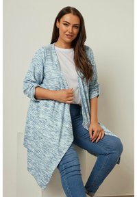 Ljusblå, texturerad kardiagan med öppen front och asymmetrisk nederkant. Bärs över en vit t-shirt och matchas med blå jeans.