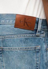 Nærbilde av baklommen på blå jeans i denim med brun Calvin Klein skinnlogo og hvit ribbestrikket skjorte stukket inn.