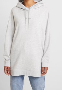 Jasnoszara, oversize bluza z kapturem, z kieszenią typu kangur i sznurkiem przy kapturze. Posiada minimalistyczne logo na klatce piersiowej. Miękka tekstura, casualowy design.