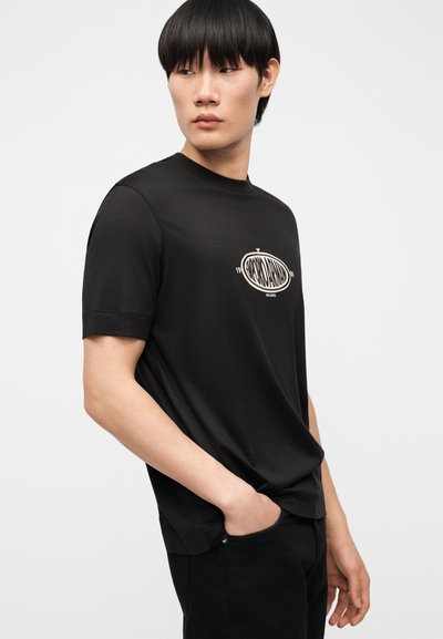 Νέος άνδρας με μαύρα, ίσια μαλλιά, που φοράει μαύρο T-shirt με κοντό μανίκι και λευκό οβάλ λογότυπο στο στήθος, στέκεται με το ένα χέρι στην τσέπη.