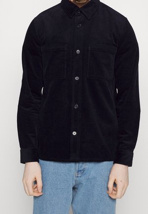 Homme portant une chemise noire en velours côtelé boutonnée avec deux poches poitrine et un jean bleu clair sur un fond blanc uni.