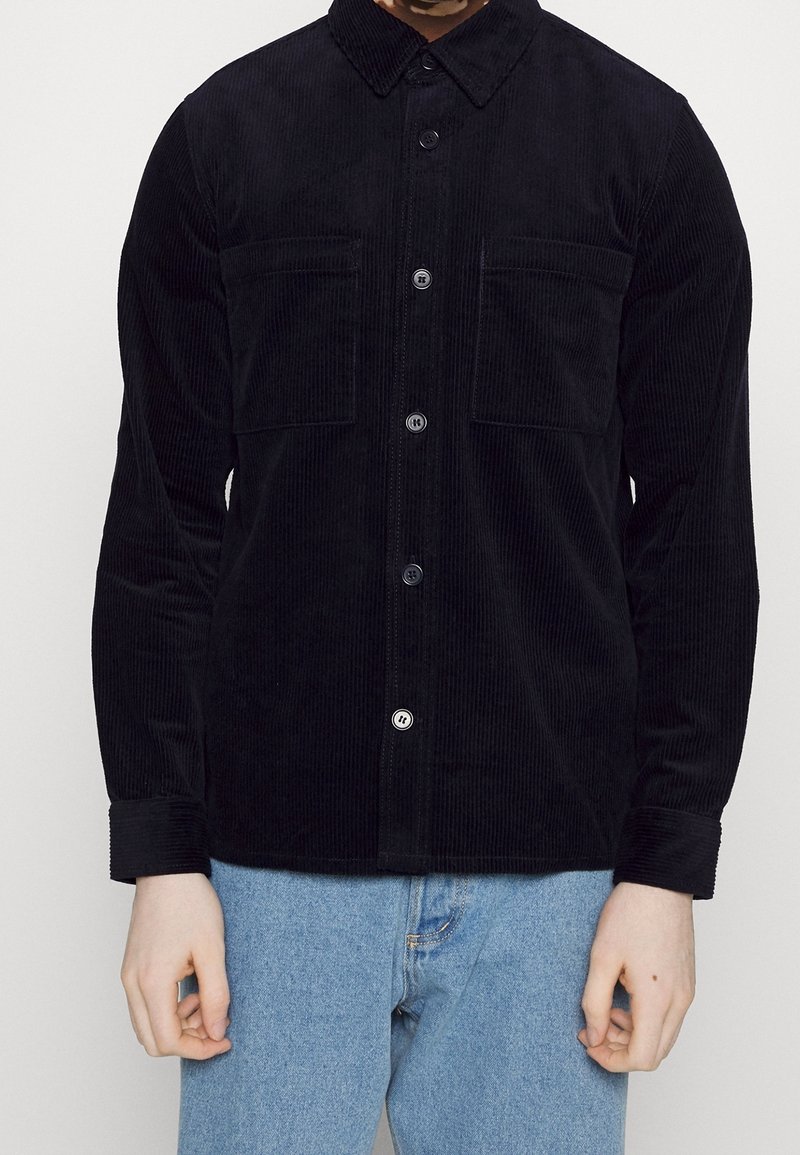 Homme portant une chemise noire en velours côtelé boutonnée avec deux poches poitrine et un jean bleu clair sur un fond blanc uni.