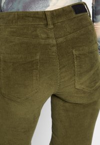 Close-up van olijfgroene corduroy broek met achterzakken en een zwart merklabel op de tailleband, gedragen met een patroon top.