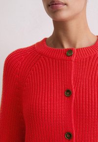 Czerwona dziergana kardiganka z okrągłym dekoltem, z trzema ciemnymi guzikami z przodu. Tekstura jest prążkowana, o średniej wadze.