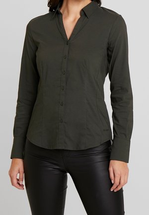 Femme portant une chemise ajustée à manches longues, boutonnée, de couleur vert foncé, et un pantalon en cuir noir sur un fond clair uni.