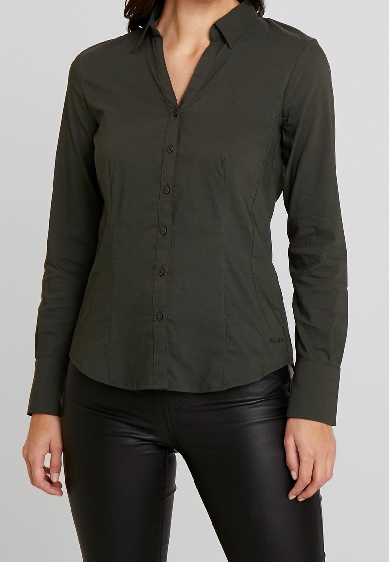 Femme portant une chemise ajustée à manches longues, boutonnée, de couleur vert foncé, et un pantalon en cuir noir sur un fond clair uni.