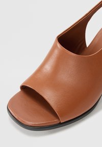 Sandalia de cuero marrón de punta abierta con una correa ancha y una silueta elegante, que presenta una textura suave y una suela negra baja.