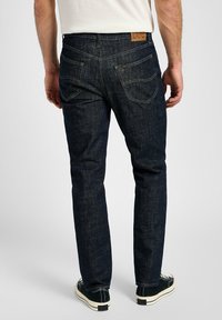Vaqueros de mezclilla azul oscuro con corte slim, que cuentan con bolsillos traseros y una etiqueta de marca de cuero marrón. El material tiene una textura suave.