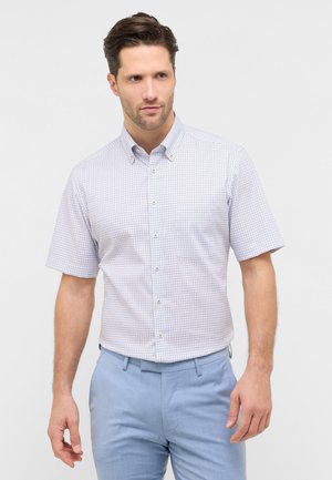 Uomo che indossa una camicia bianca a maniche corte con piccoli quadretti blu e rossi e pantaloni azzurri, in piedi davanti a uno sfondo semplice.