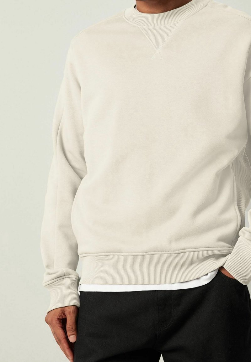 Sudadera de color beige claro con cuello redondo acanalado y mangas con puños. Tejido suave, ajuste relajado, combinada con una camiseta blanca y pantalones negros.