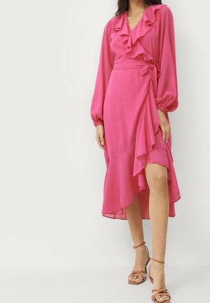 Femme portant une robe portefeuille rose à manches longues avec des volants et un lien sur le côté, associée à des sandales à talons hauts beiges à brides.