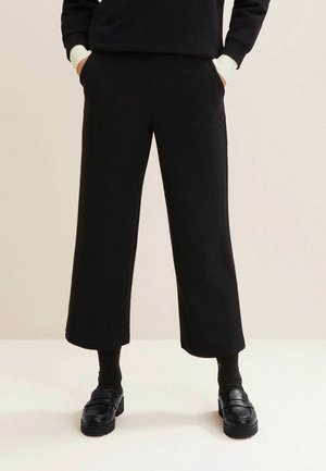 Person trägt schwarze, weite, kurze Hose, schwarze Socken, schwarze Loafers und ein schwarzes Langarmoberteil mit weißen Manschetten, die Hände in den Taschen.