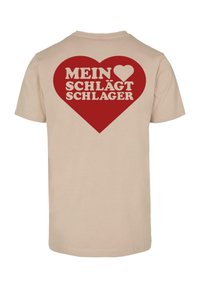 Beige medvilninė marškinėliai su dideliu raudonu širdies grafiniu paveikslu ir balta užrašu "MEIN ❤️ SCHLÄGT SCHLAGER" ant nugaros.
