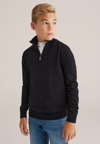 Blonds zēns, kas ģērbts melnā pus-izvilkuma džemperī virs balta krekla un ziliem džinsiem, skatās pa kreisi uz vienkārša fona.