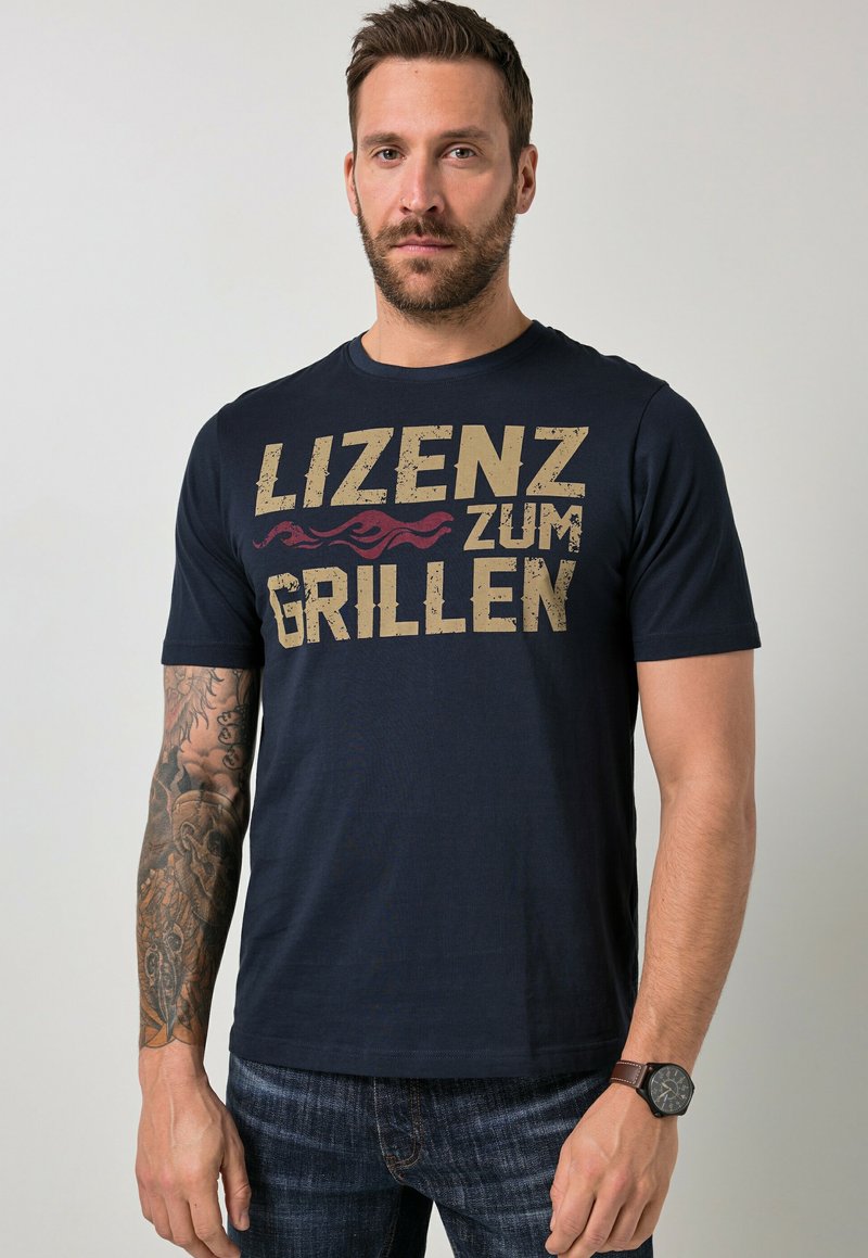 Mann mit kurzem braunem Haar und Bart, trägt ein schwarzes T-Shirt mit dem Schriftzug "Lizenz zum Grillen" und Flammengrafik, dazu Jeans und eine Armbanduhr.