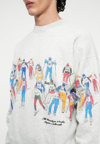Szary sweatshirt z kolorowymi figurami narciarzy nadrukowanymi na niebiesko, czerwono, żółto i czarno. Napis brzmi: „The Mountain Nordic Aspen, Colorado.”