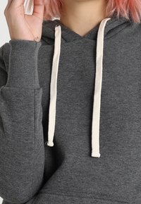 Persona che indossa un felpa grigia con cordini bianchi, tiene la mano vicino al viso, mostrando capelli rosa e un piccolo tatuaggio sul polso.