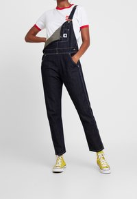 Pantalones de mezclilla con tirantes ajustables, color azul oscuro, costuras en contraste, bolsillos frontales y una parche gris en el peto. Combinados con una camiseta blanca.