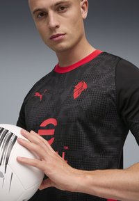 Sort og rød kortærmet trøje med et løve-logo, struktureret mønster og fed skrift. Model holder en hvid fodbold.