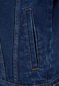 Gros plan d'une poche de veste en denim bleu foncé avec ouverture verticale et détail de couture orange le long des coutures et des bords.