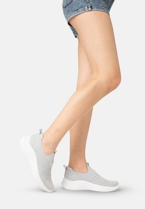 Ben iført grå slip-on strikkede sneakers med hvide såler, iført denimshorts, placeret mod en ensartet lys baggrund.