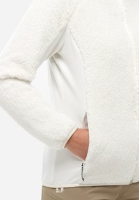 Main dans la poche zippée latérale d'une veste en polaire blanche avec un tissu doux et un pantalon beige portant le logo Millet.