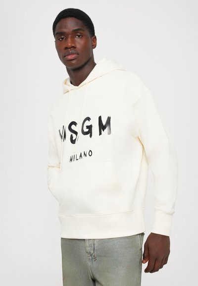 Uomo giovane che indossa una felpa MSGM Milano color avorio e jeans azzurri chiari, in piedi contro uno sfondo bianco semplice.
