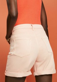 Shorts de mezclilla melocotón con dobladillo enrollado, que cuentan con dos bolsillos traseros y una textura suave. El diseño incluye detalles de costura sutiles.